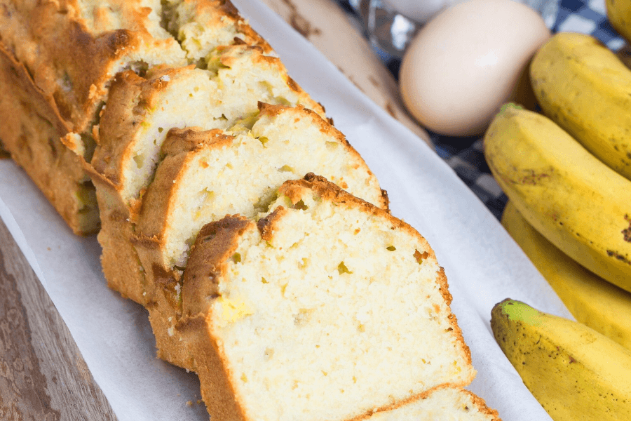 A freshly baked loaf of 4-ingredient banana bread on a wooden cutting board, surrounded by ripe bananas and a knife for slicing.