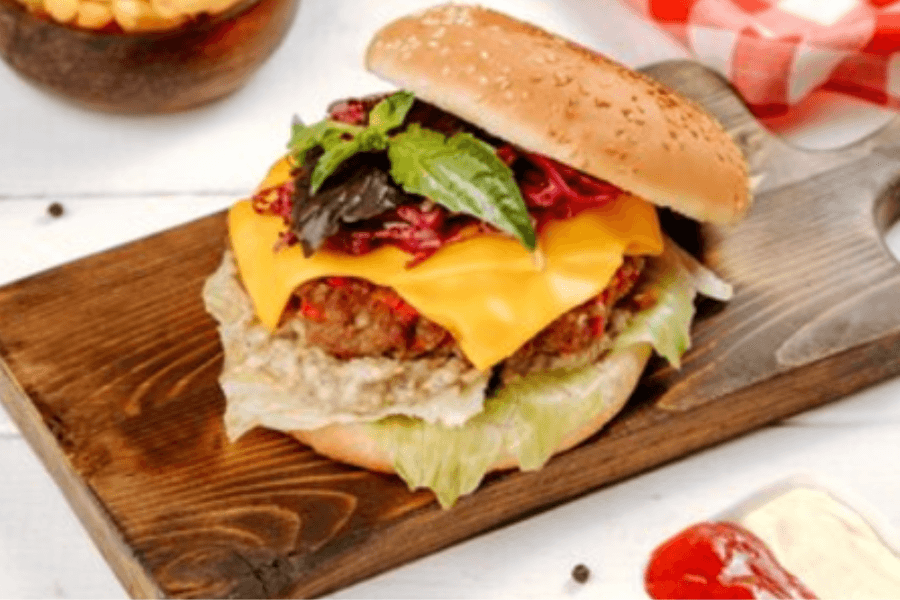 A gourmet burger with a sesame seed bun, a juicy beef patty topped with melted cheddar cheese, fresh lettuce, a dollop of creamy sauce, vibrant red cabbage slaw, and garnished with basil leaves, served on a wooden board.